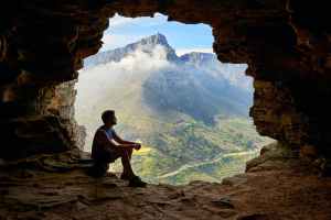 Man sitting in the mouth of a cave with open sky and mountain showing beyond the cave