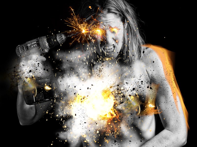 A person that is exploding with sparks from the inside out holds a cordless drill to their own temple, which is also producing sparks.