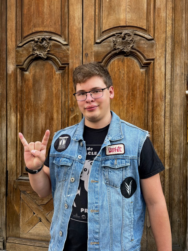 White teenager standing in front of a large, old wooden door wearing a denim punk battle jacket covered in band patches.