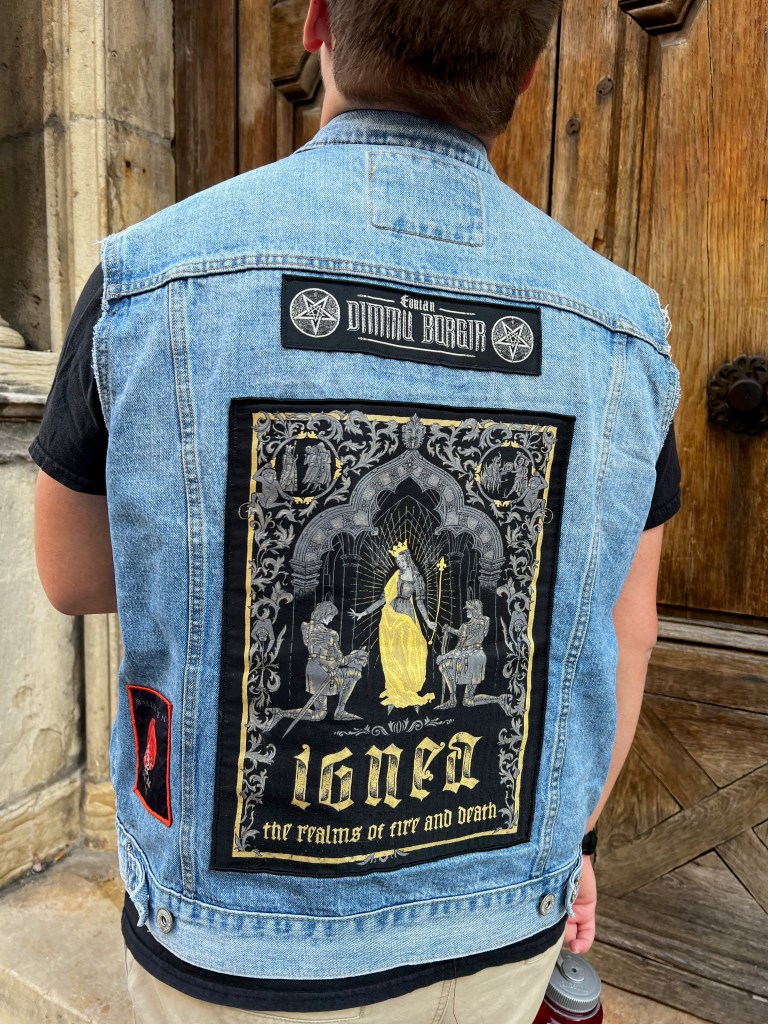 White teenager standing in front of a large, old wooden door wearing a denim punk battle jacket covered in band patches.