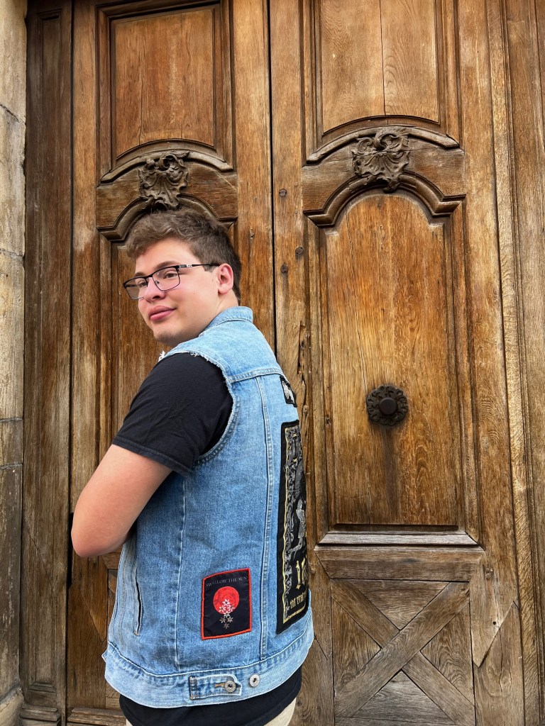 White teenager standing in front of a large, old wooden door wearing a denim punk battle jacket covered in band patches.