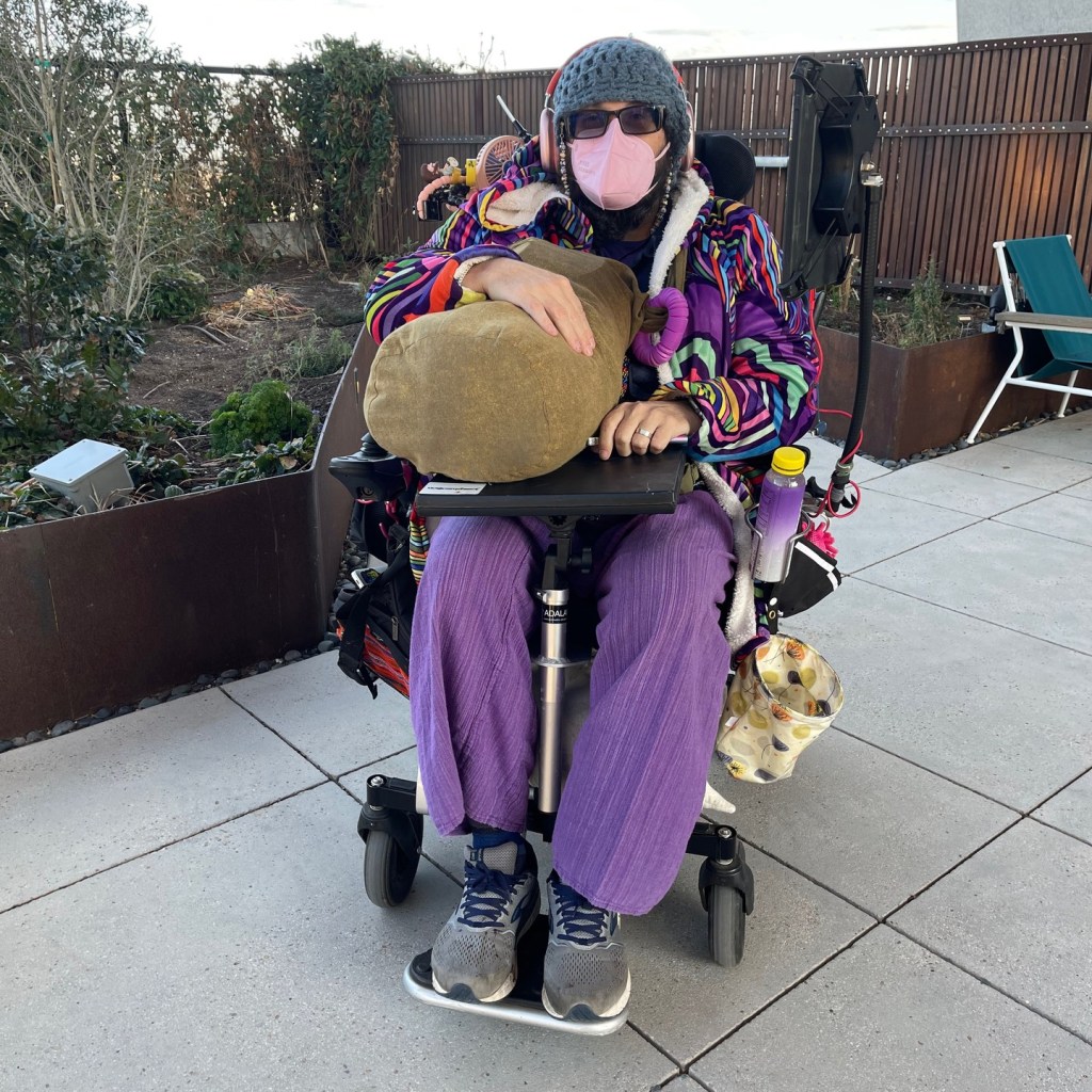A middle aged white man dressed in purple pants and bright colored coat and wearing sunglasses, beanie hat, and a face mask sits in a power wheelchair