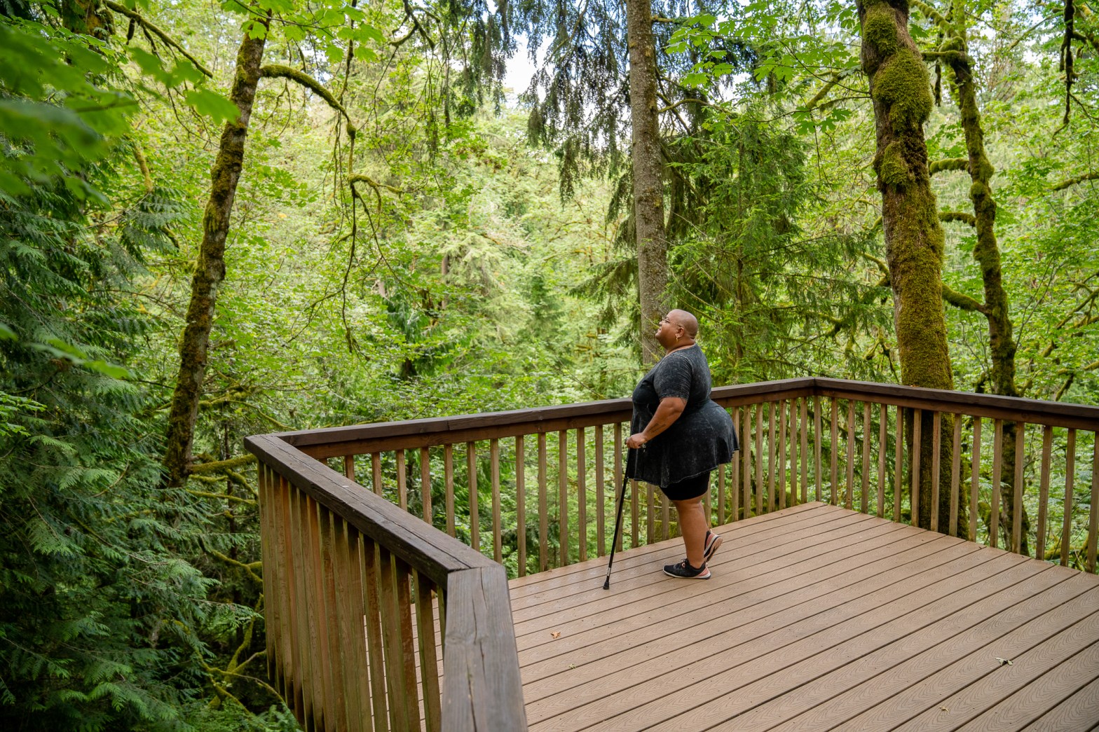 A Black non-binary hiker stands on a wooden deck with their cane, looking out into the surrounding forest. They have a shaved head and wear glasses, a peplum shirt, shorts, and tennis shoes.
