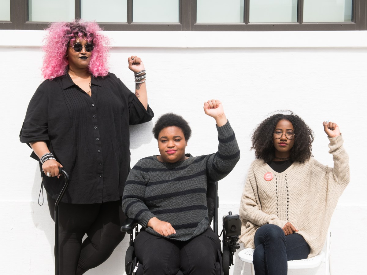 Torso level photo of three Black and disabled folx (a non-binary person holding a cane, a non-binary person in a power wheelchair, and a femme on a folding chair) raising their fists on the sidewalk in front of a white wall.