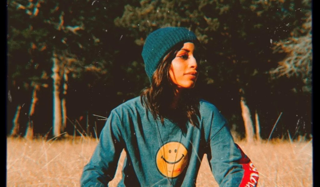 אישה ארוכת שיער חומה עם כובע כפה וחולצת סמיילי מביטה מהמצלמה