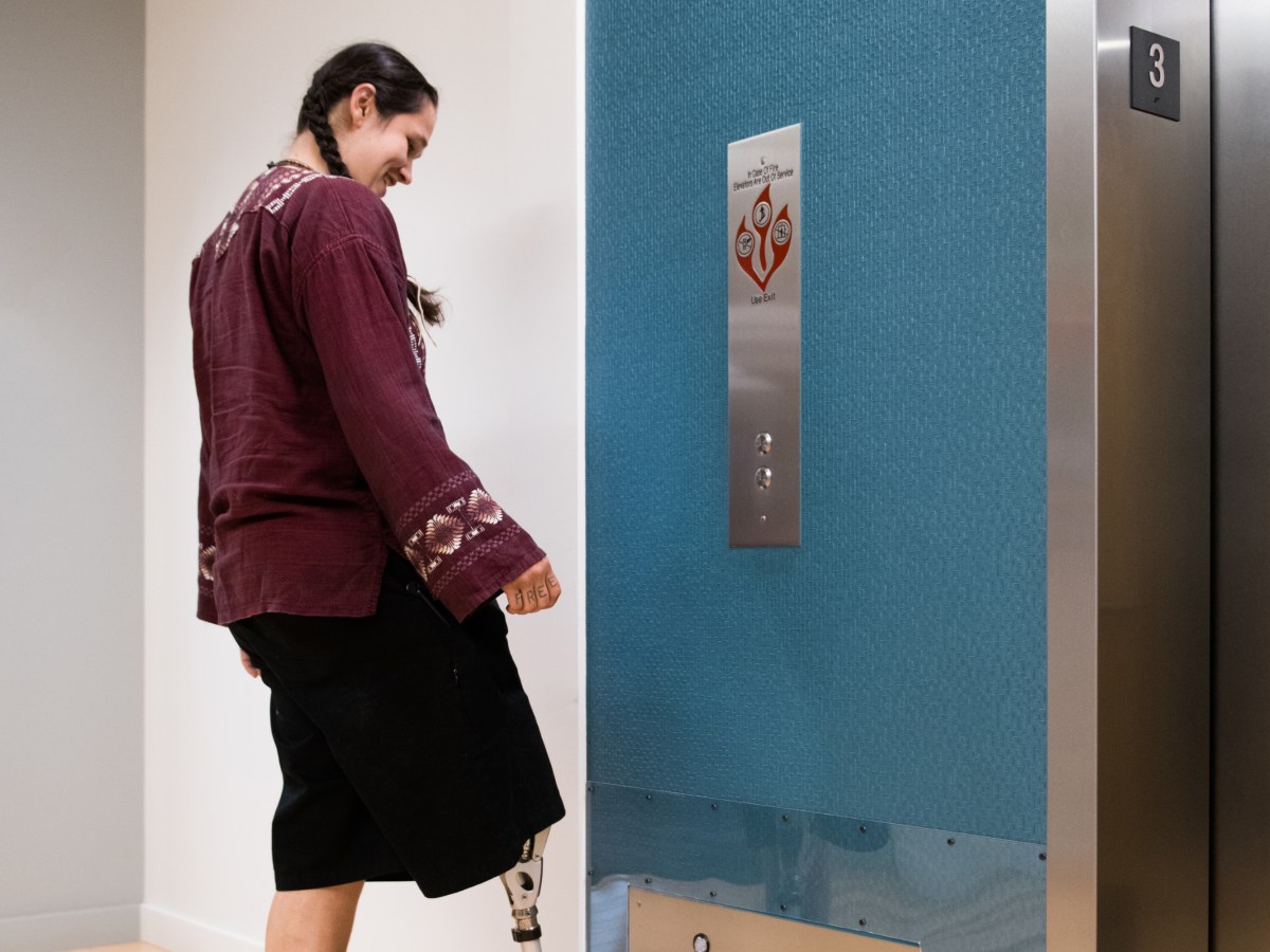 An Indigenous Two-Spirit person calls the elevator up by tapping the floor button with their prosthetic leg.