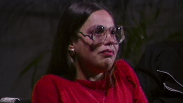 A young white woman with long dark hair and round glasses speaks into a microphone