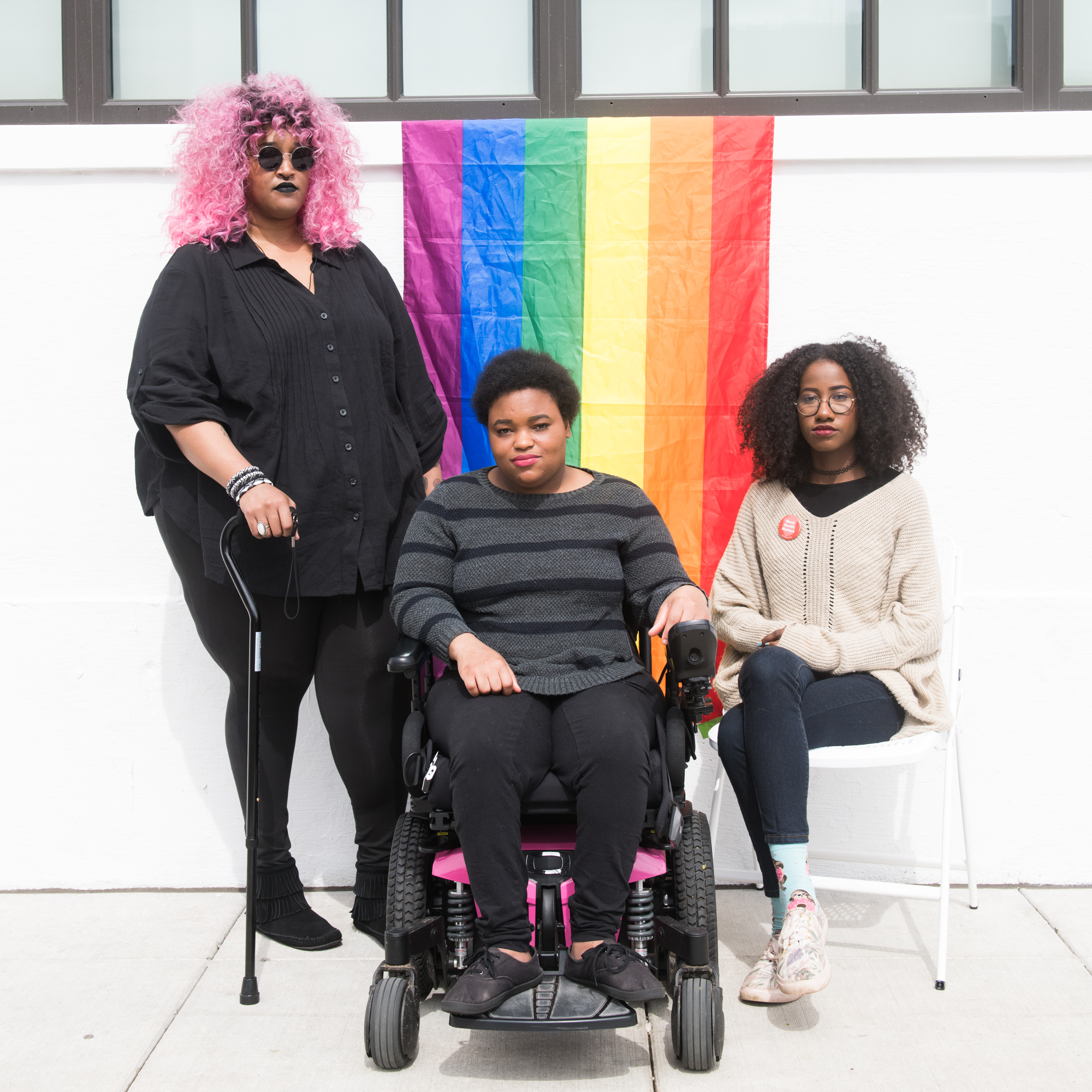 Three Black and disabled folx (a non-binary person holding a cane, a non-binary person sitting in a power wheelchair, and a femme sitting in a chair) look seriously at the camera while a rainbow pride flag drapes on the wall behind them.

