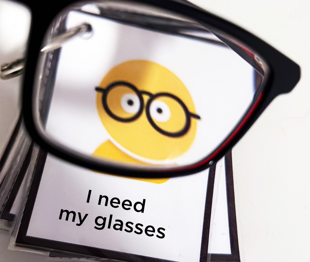Photo of a communication card featuring a yellow figure wearing black glasses. The card is viewed through the lens of an actual pair of black eyeglasses.