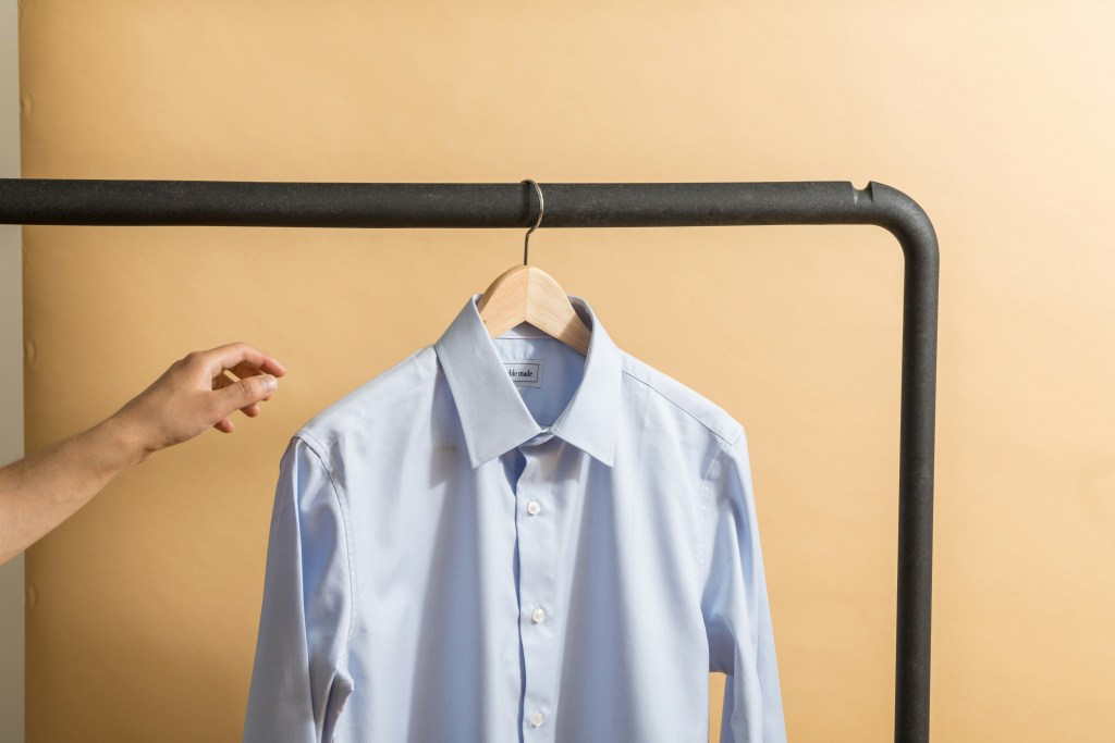A hand reaches out towards a white dress shirt on a wooden hanger on a clothes rail