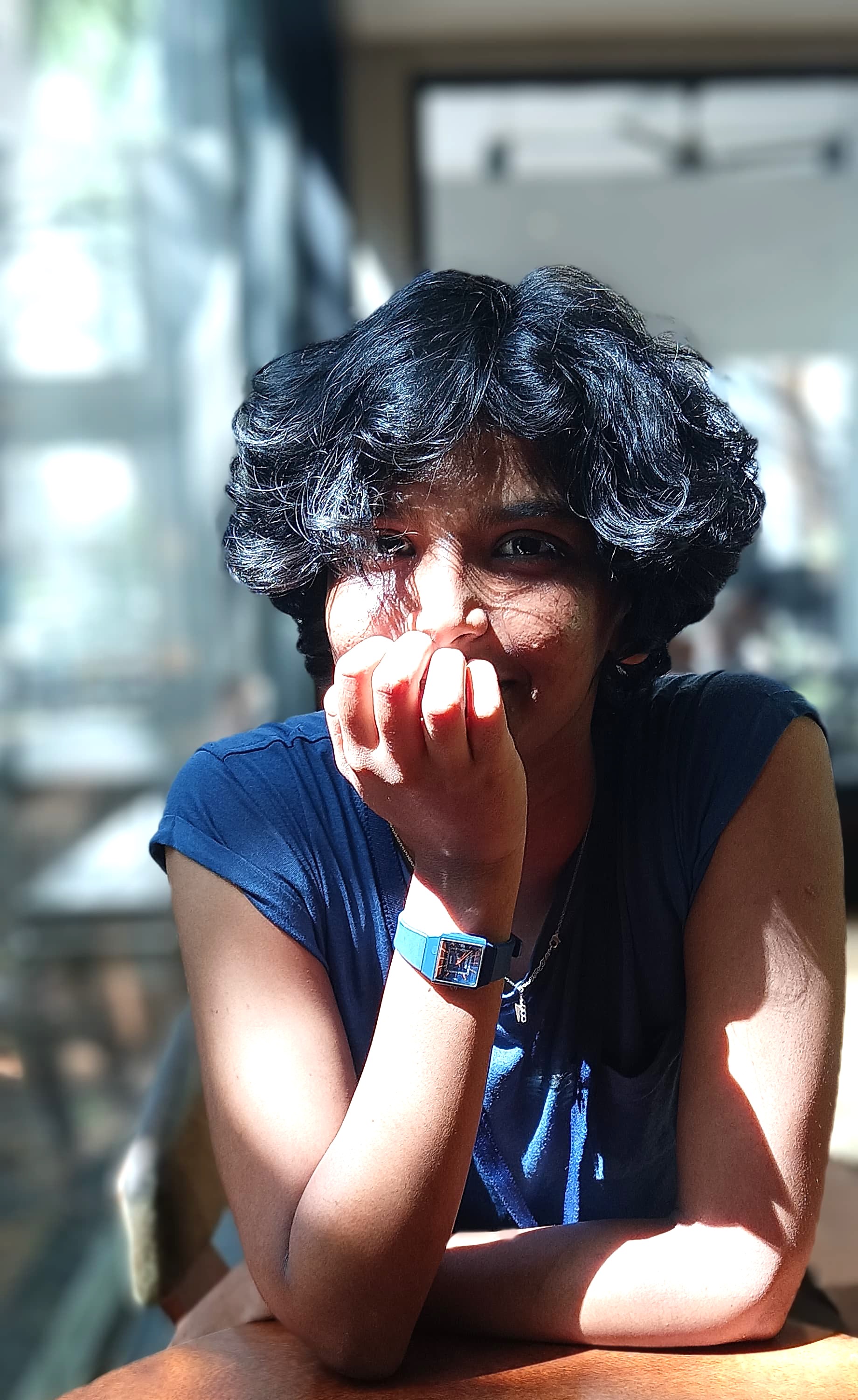 A brown-skinned woman with short black hair smiles behind fingers curled at her moment. She's wearing a short sleeve blue shirt and sitting  at a table in a sunbeam.