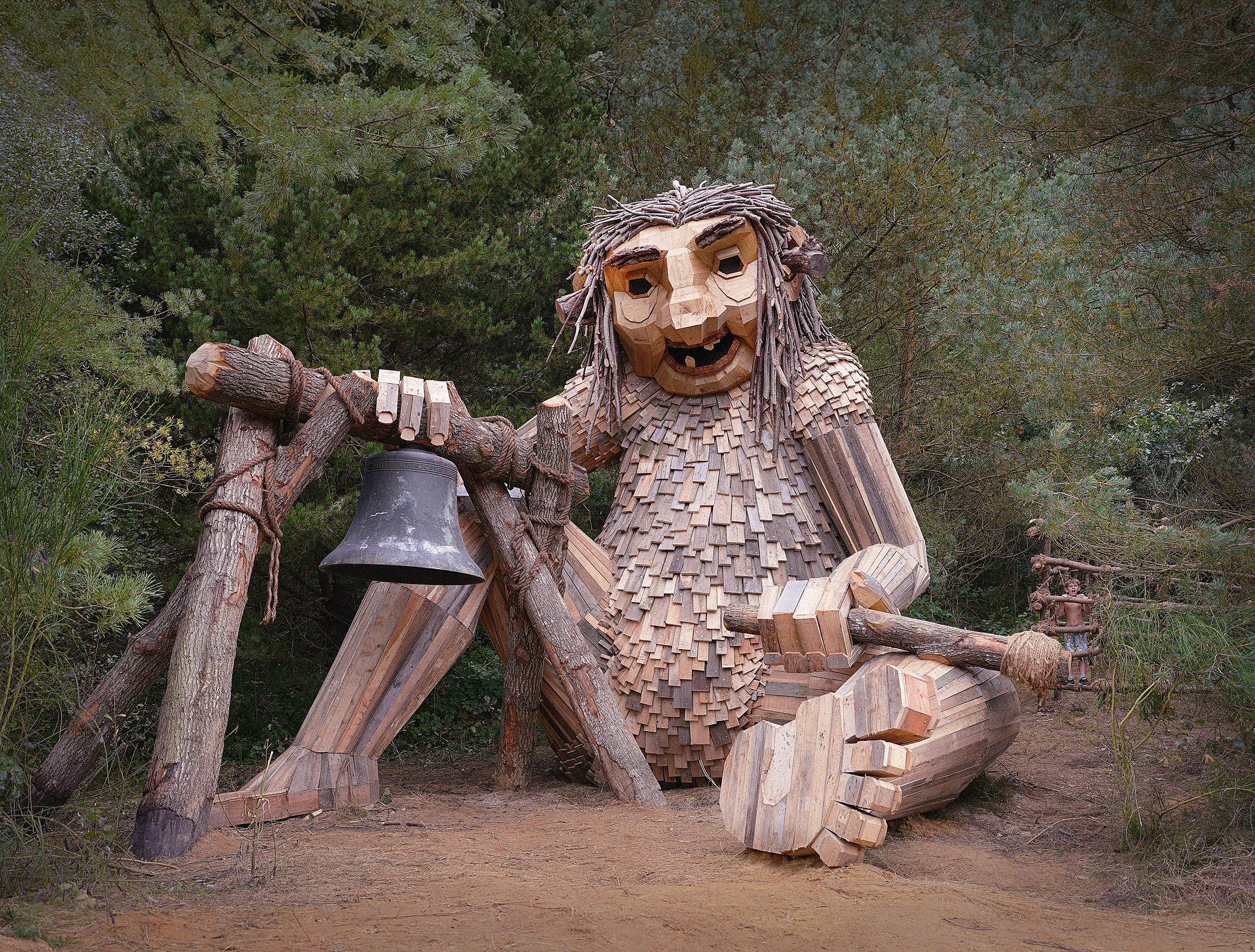 A large wooden troll sculpture by Thomas Dambo sits in a forest clearing, constructed from reclaimed lumber, branches, and wood shingles. The troll has a wide, expressive face with gap-toothed mouth and twig-like hair, and holds a large iron bell suspended from a wooden frame in one hand. Its body is covered in overlapping wooden slats resembling scales or feathers. A small human figure is visible in the background to the right, emphasizing the sculpture's massive scale. Sandy ground, pine trees.