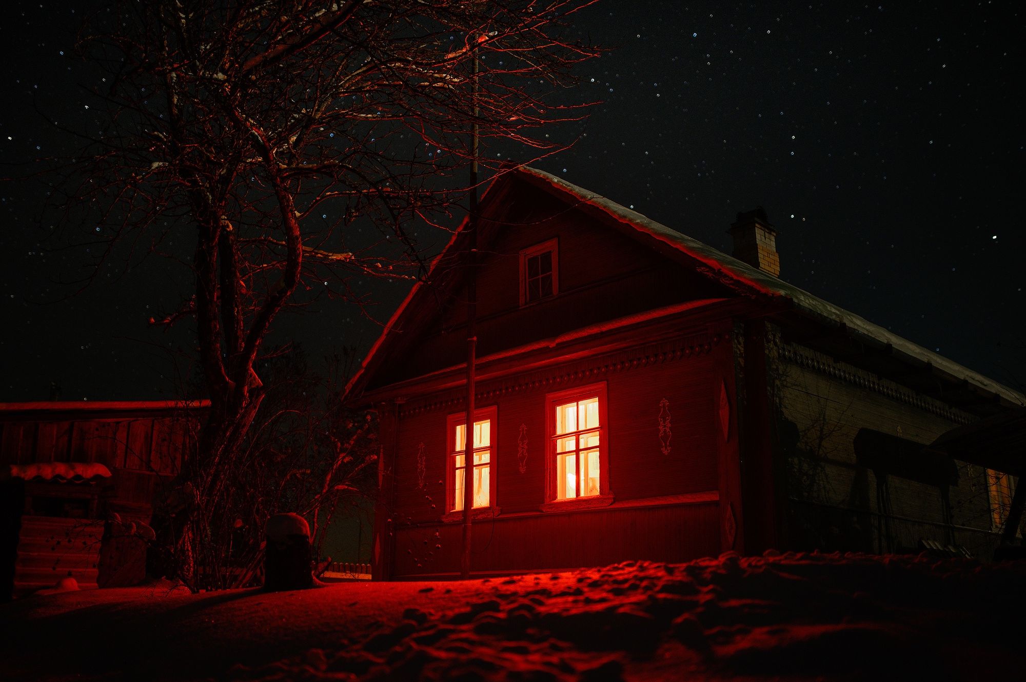 A wooden house at night under a starry sky, washed in red light, with warm yellow light glowing in two windows. A bare tree stands beside it, and snow covers the ground.