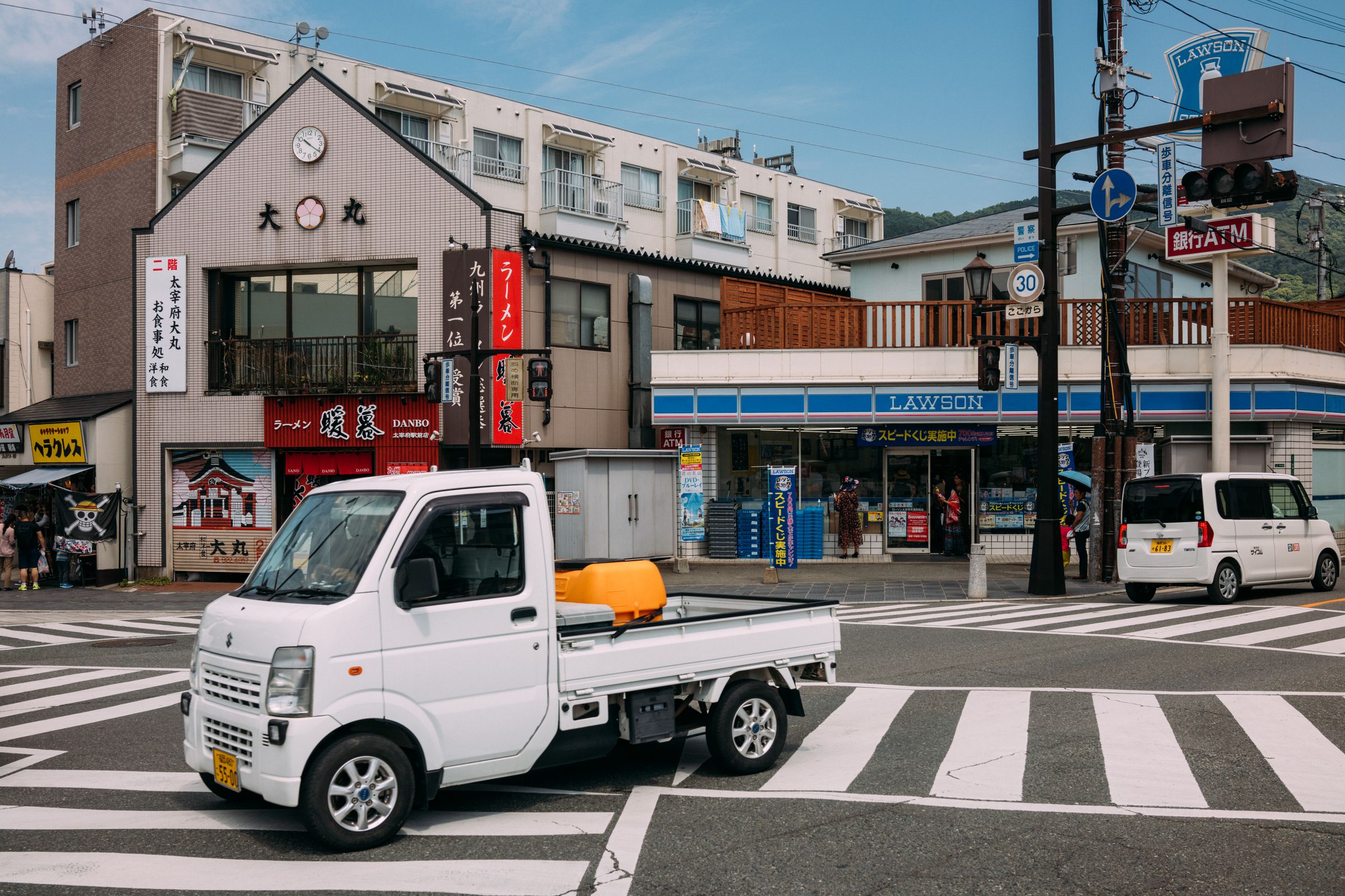 A small white kei truck with an orange container in its bed crosses a zebra-striped intersection in a Japanese town, in front of a ramen shop and a Lawson convenience store.