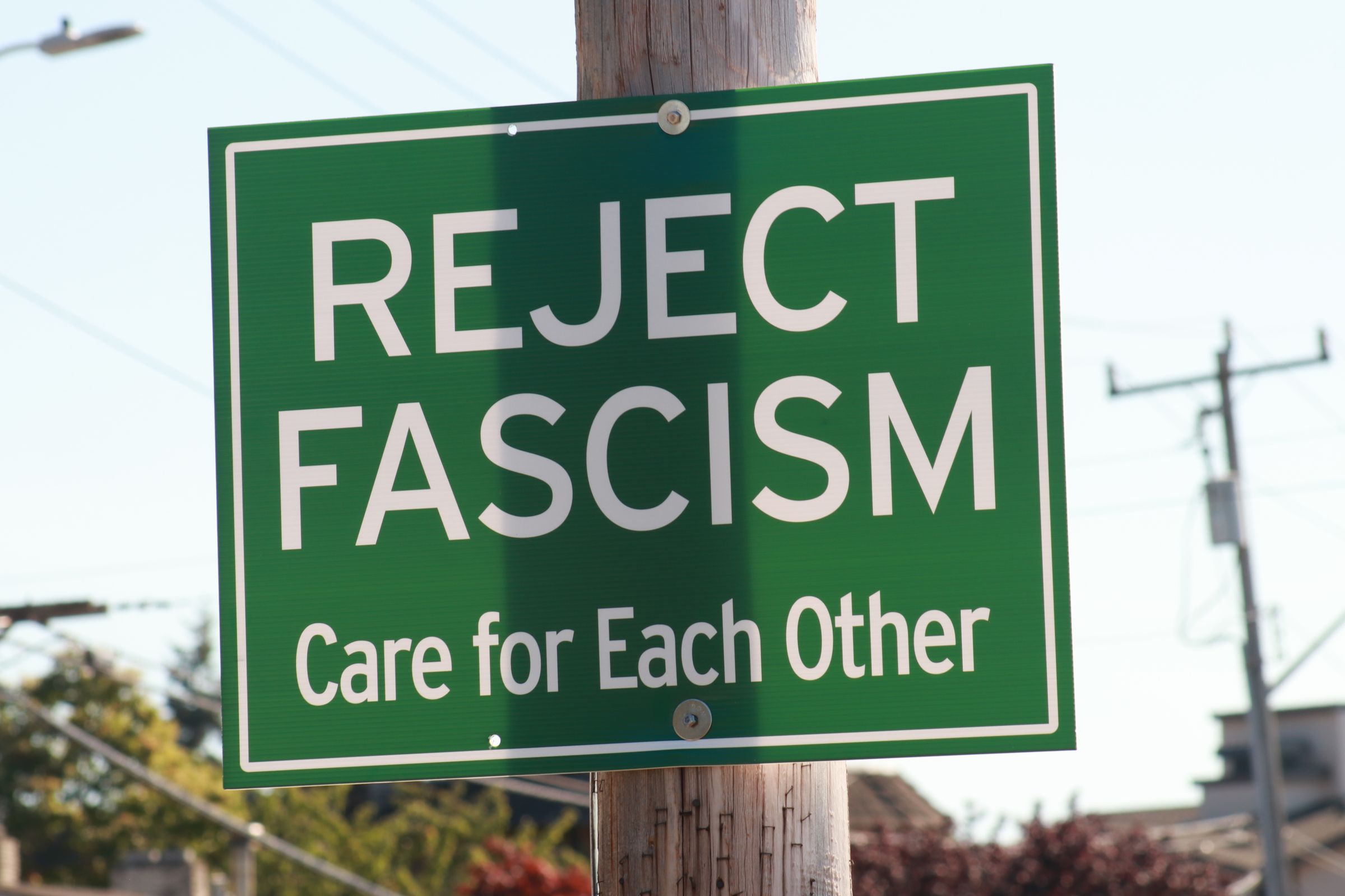 A green street sign bolted to a wooden utility pole reads, in white letters: “Reject Fascism. Care for Each Other.” Power lines, a second pole, and rooftops sit behind it under a pale sky.