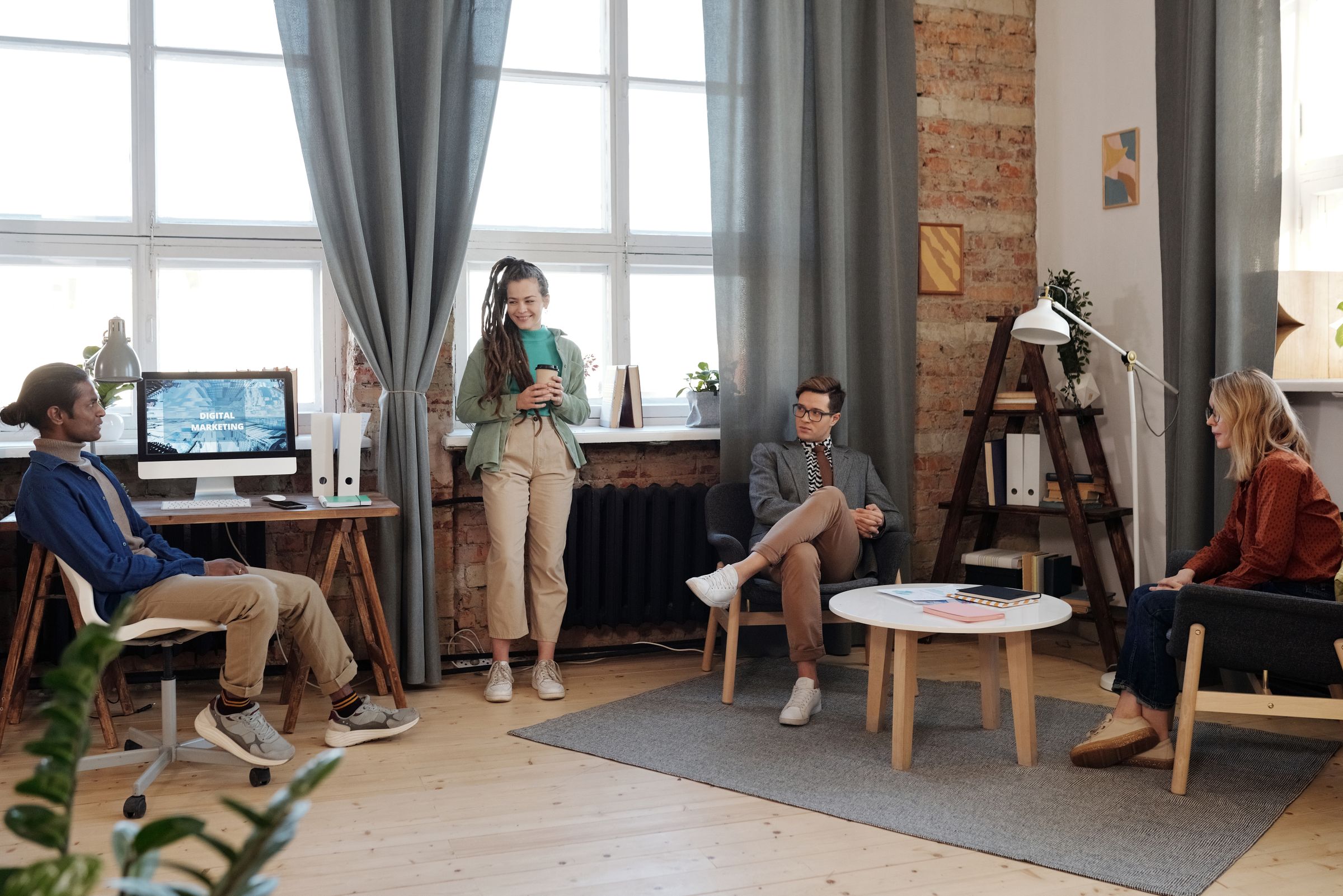 Four people gathered in a bright room with tall windows, grey curtains, and a bare brick wall. One sits at a desk, turned away from the computer to face the others. One stands by the window holding a cup and smiling. Two sit in armchairs around a small round table, listening.